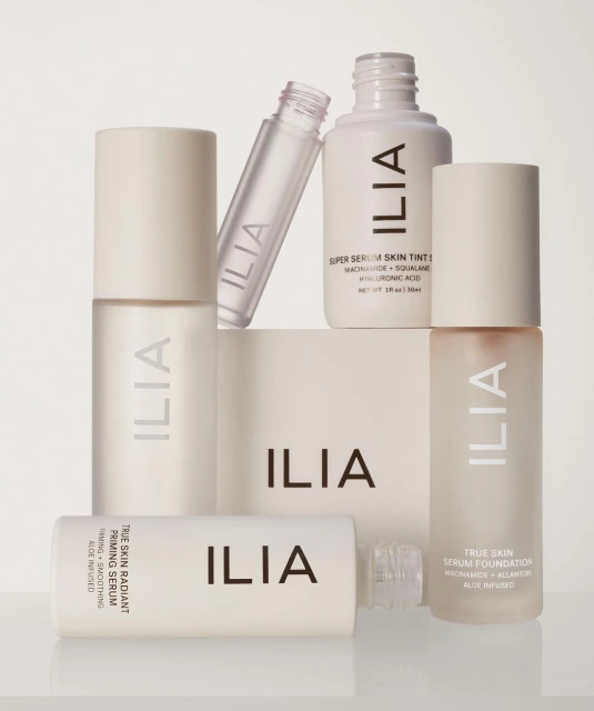 Several bottles and containers of Ilia beauty and skincare products are arranged together against a soft, neutral background.