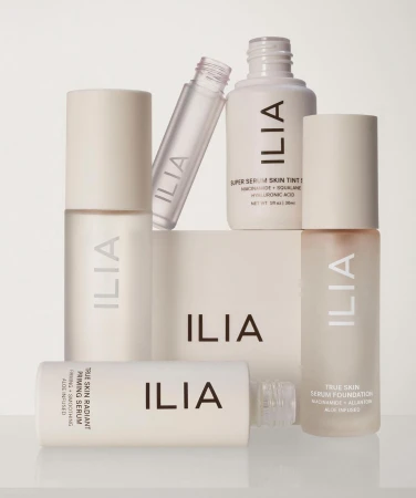 Several bottles and containers of Ilia beauty and skincare products are arranged together against a soft, neutral background.