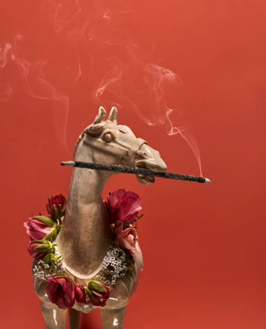 Statue of a horse holding an incense stick with smoke. Statue is decorated with a wreath of roses against a vibrant coral background.