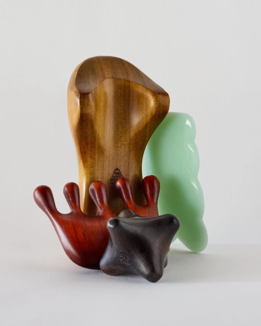Several wooden gua sha massage tools on a white surface including a brown paddle, small dark tool, and green clear tool.