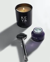 Product still life of a burning candle, facial roller, and skincare jar on a white background by John Von Pamer.