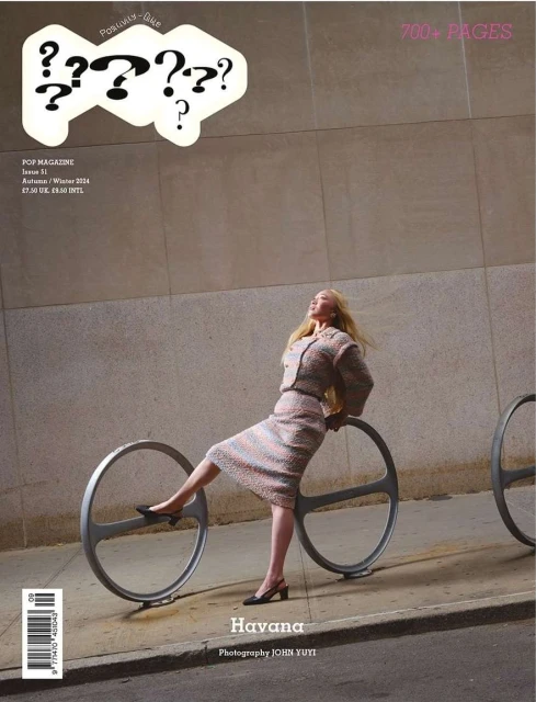 Magazine cover featuring a blonde woman in a patterned suit leaning against a round metal bike rack, photographed in an urban setting.