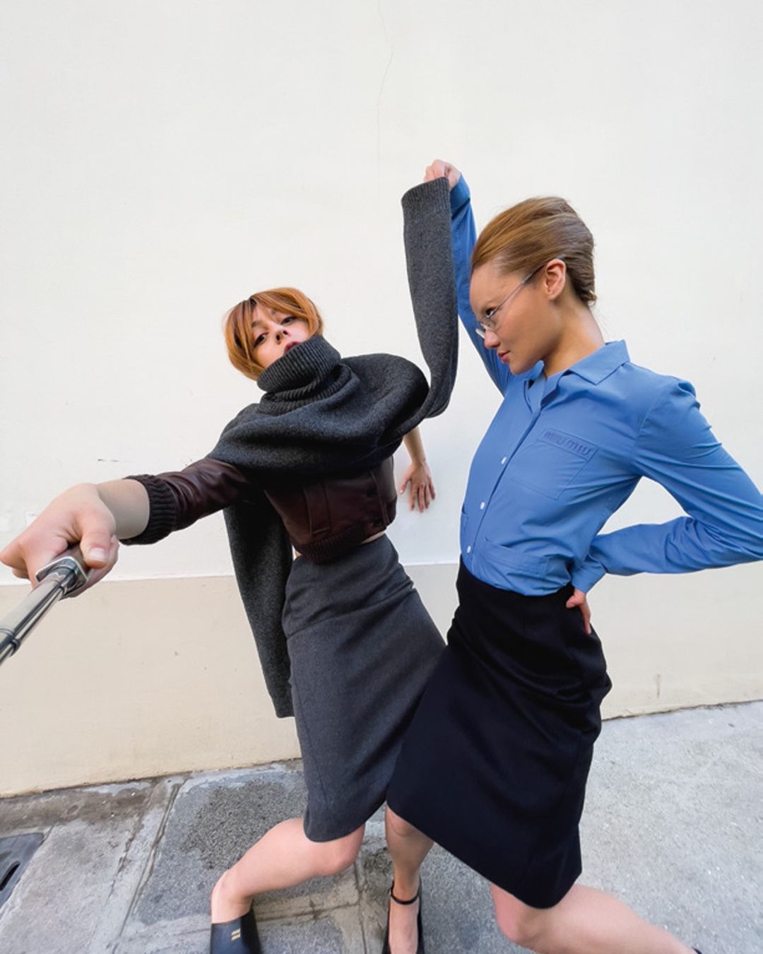 Two adult women in skirts and blouses strike poses in an urban setting, one using a selfie stick, under bright, natural lighting.