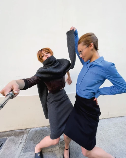 Two adult women in skirts and blouses strike poses in an urban setting, one using a selfie stick, under bright, natural lighting.