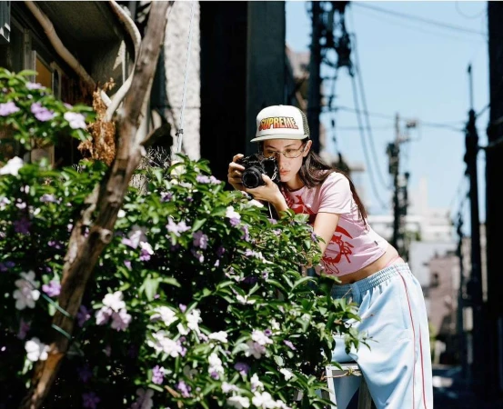 A young adult woman with glasses and a cap takes a photo with a camera next to a flowering bush in an alleyway on a sunny day.