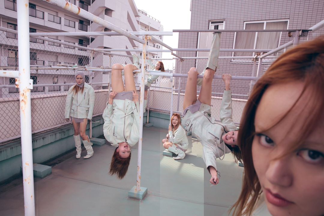 Group of young women in light-colored jackets posing playfully on a rooftop with a metal structure against a backdrop of buildings.