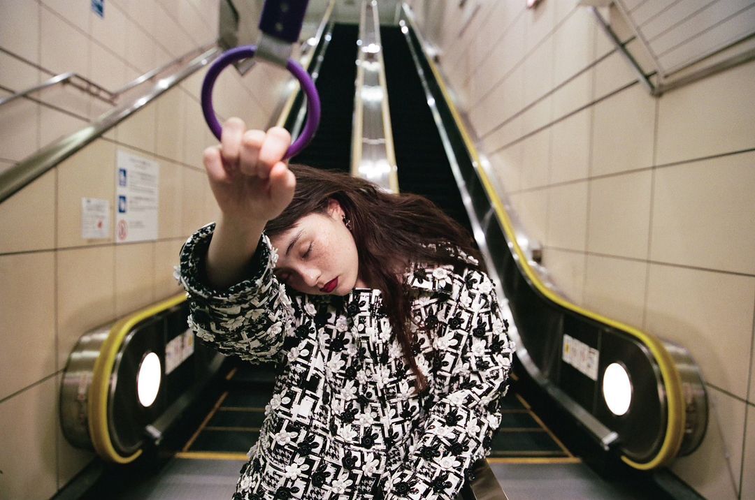 Woman with dark hair leans on an escalator handrail inside of an underground station wearing a patterned jacket with dark lipstick.