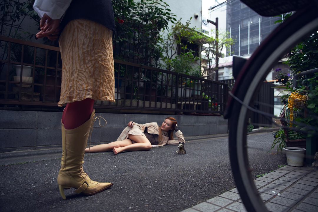 Woman in coat lying on pavement beside a toy, with another woman standing nearby and a bike wheel partially in the frame.