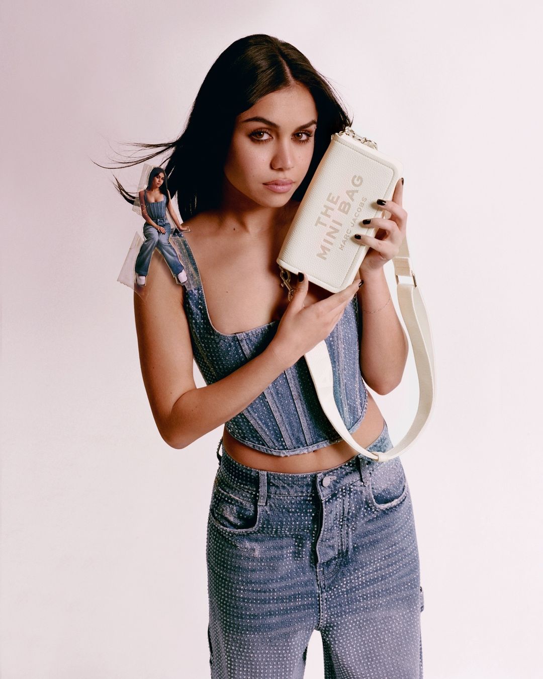 Brunette woman poses in denim corset and jeans with a white Marc Jacobs Mini Bag against a clean white background in a studio shot.