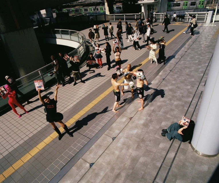 Outdoor shot captures boxers, audience on gray-tiled platform by train tracks, with colorful outfits and magazine props adding quirky flair.