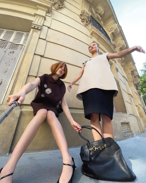 Two adult women in shift dresses pose on a sidewalk in front of a building with beige walls and shuttered windows, one holding a black handbag.