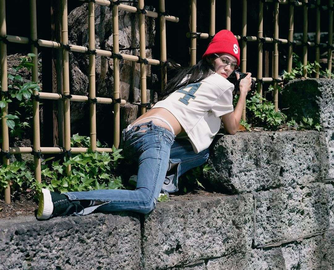 Young adult woman with red beanie poses on stone wall with camera in outdoor setting, wearing jeans and baseball shirt with bamboo fence.