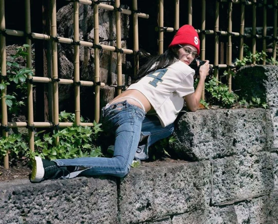 Young adult woman with red beanie poses on stone wall with camera in outdoor setting, wearing jeans and baseball shirt with bamboo fence.