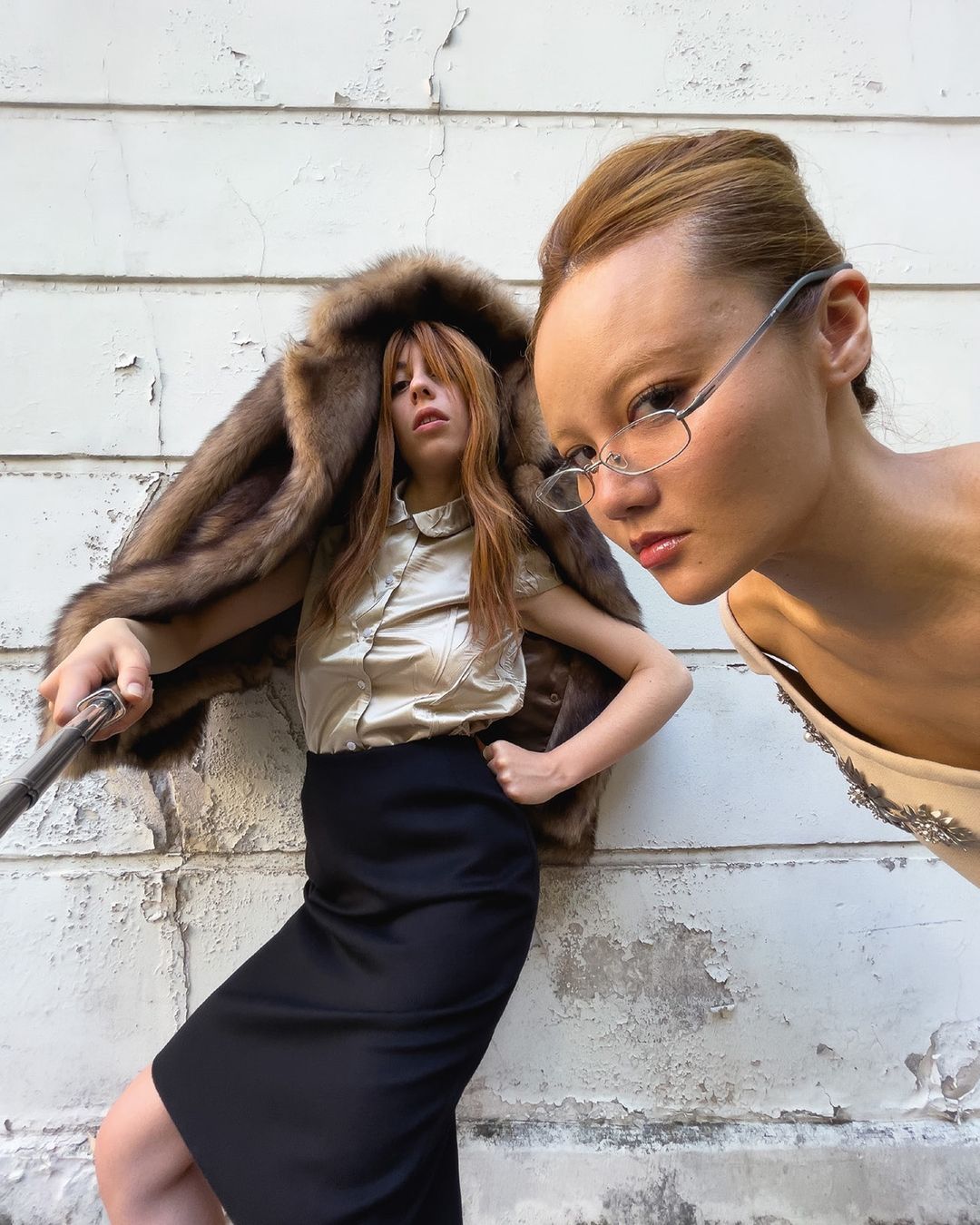 Two women pose outdoors. One woman wears glasses and a dress, while the other wears a fur coat, blouse, and skirt against a white wall.