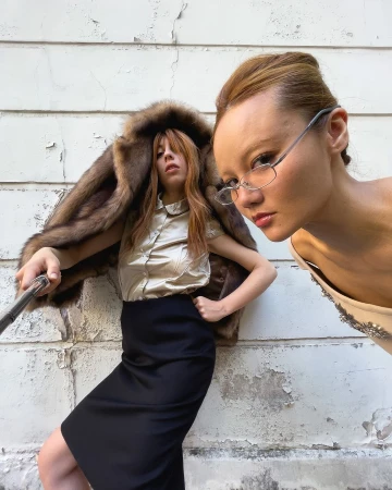 Two women pose outdoors. One woman wears glasses and a dress, while the other wears a fur coat, blouse, and skirt against a white wall.