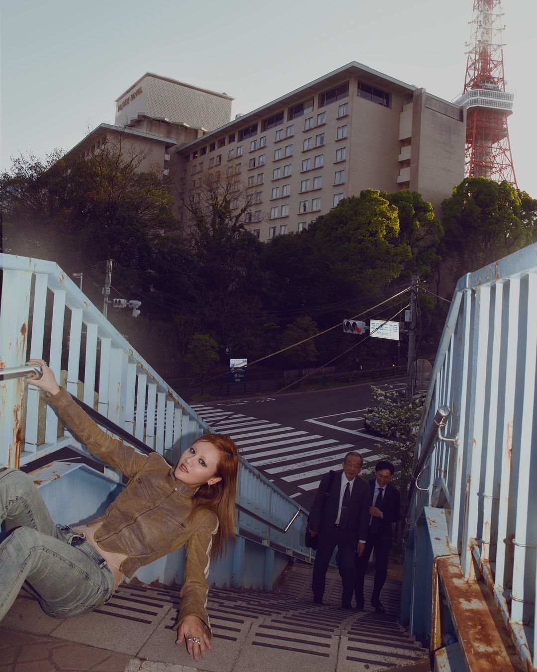 Woman in jacket and jeans poses on stairs with city tower and business people in the background on a sunny day in an urban environment.