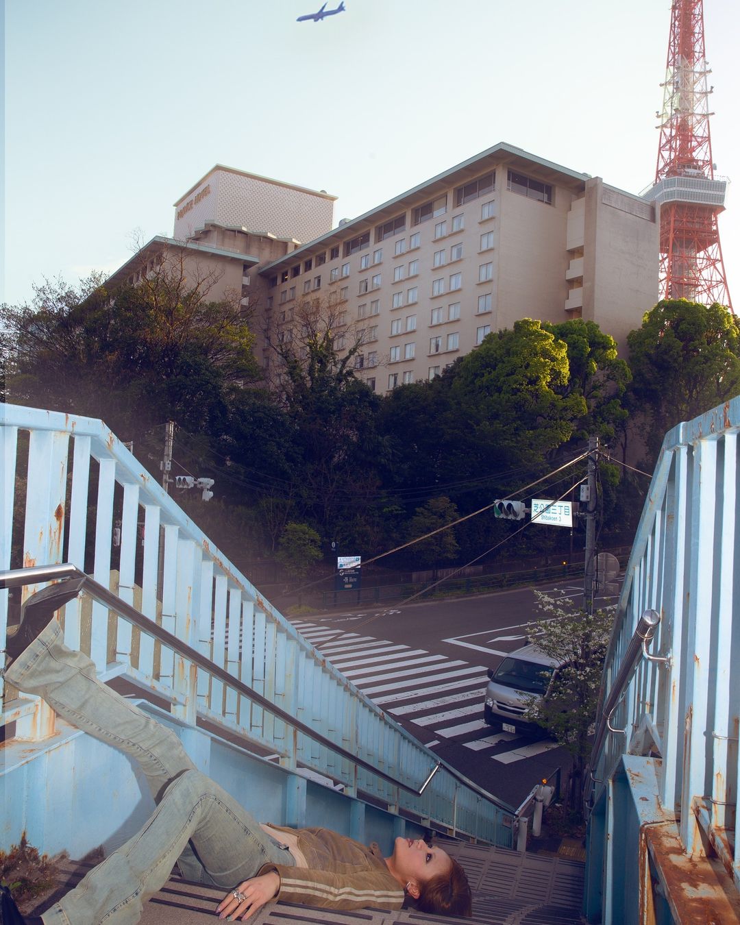 A woman lies on an outdoor staircase in an urban setting, with a tower and building in the background, beneath a blue sky and airplane.