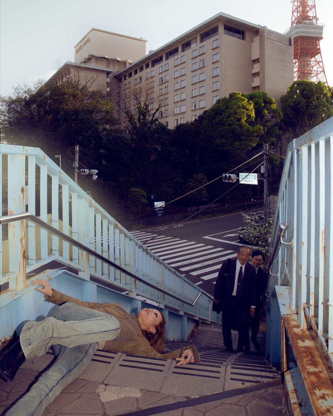 Woman laying on steps near two men in suits, with a hotel and the Tokyo Tower in the background, captured at eye-level.
