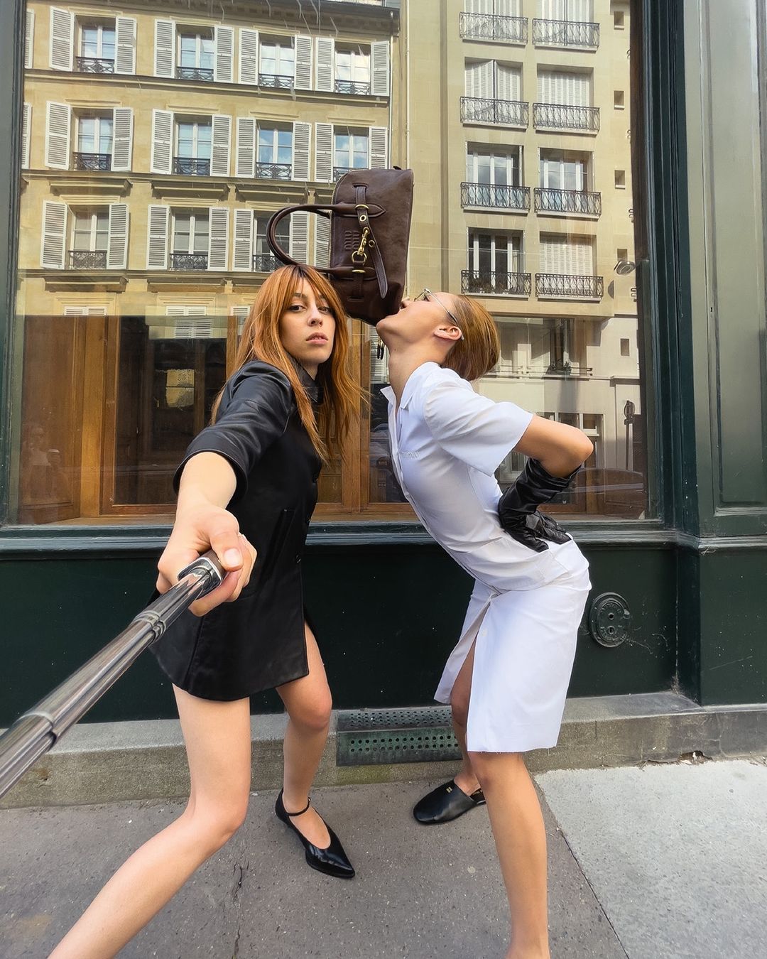 Two women pose outdoors on a city street with a brown bag balanced on one woman's head, taken with a selfie stick.
