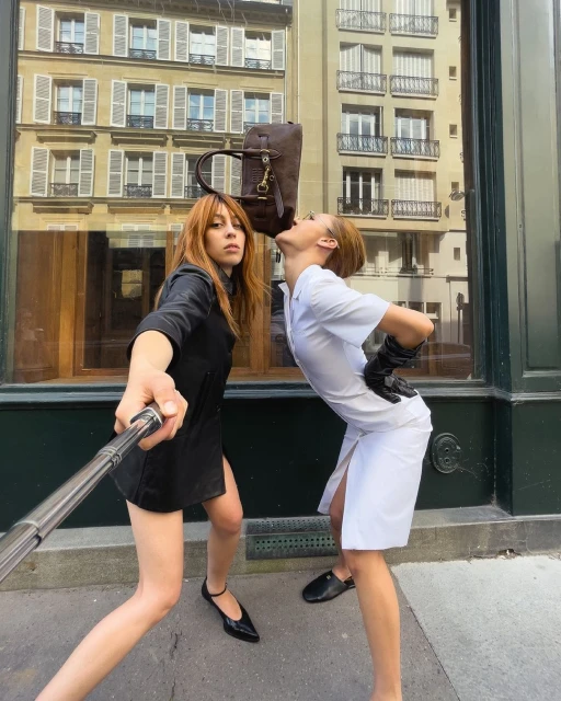 Two women pose outdoors on a city street with a brown bag balanced on one woman's head, taken with a selfie stick.