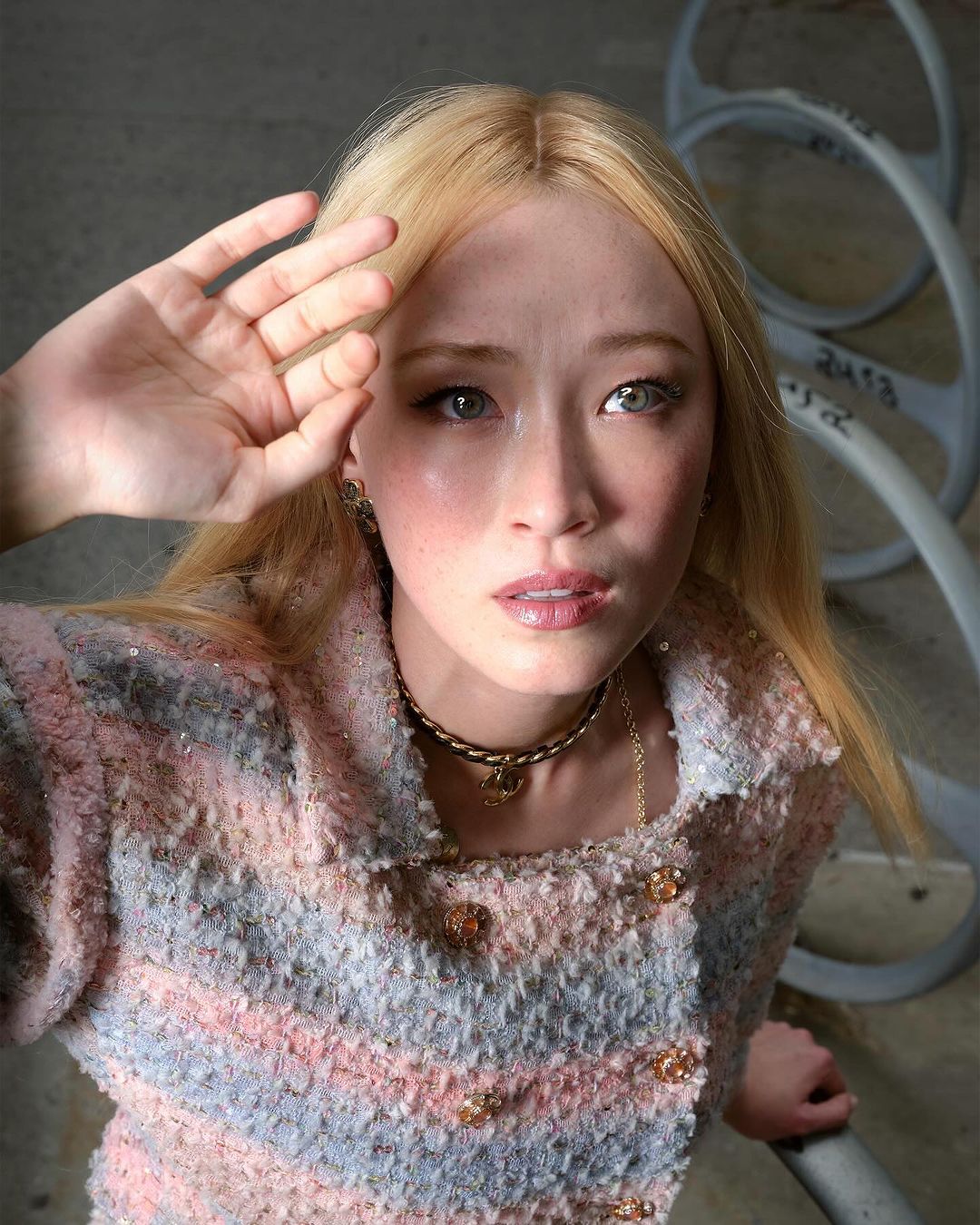 Eye-level shot of a blonde woman in a Chanel-style jacket, shielding her eyes, with concrete and metal structures visible in the background.