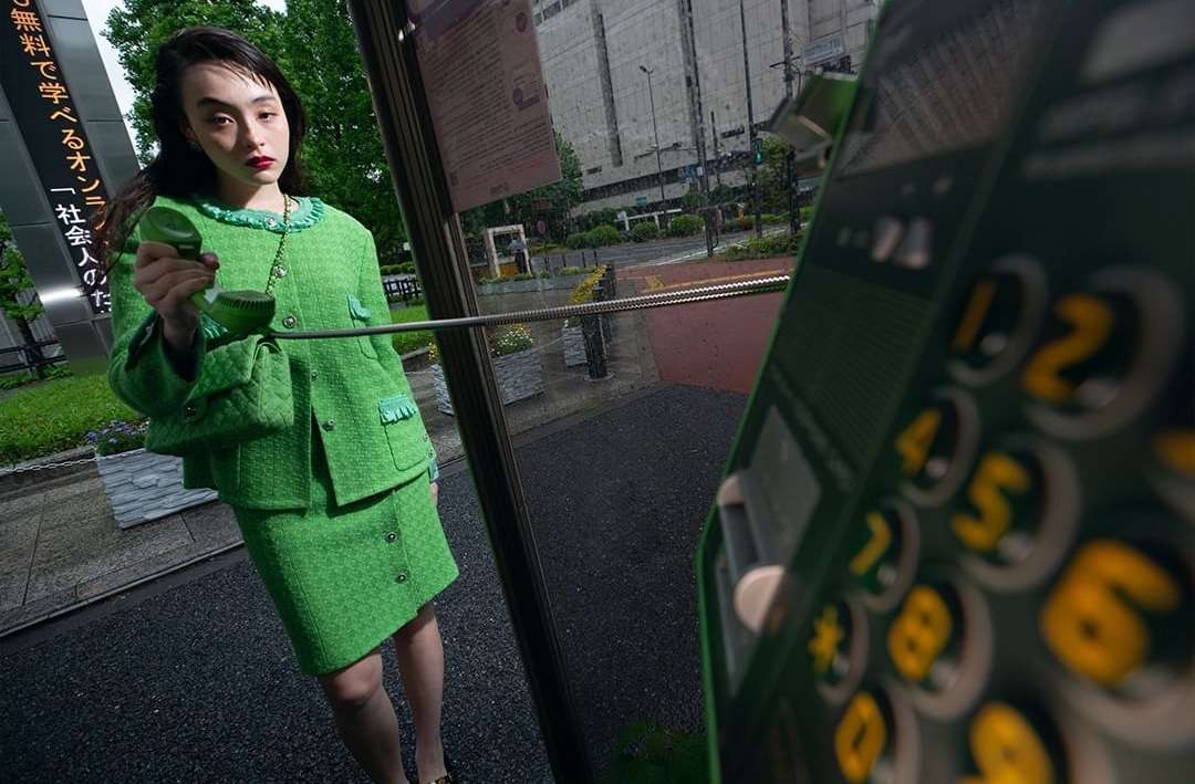 Woman in green tweed suit holds a green phone in a public phone booth on a city street with buildings in the background.
