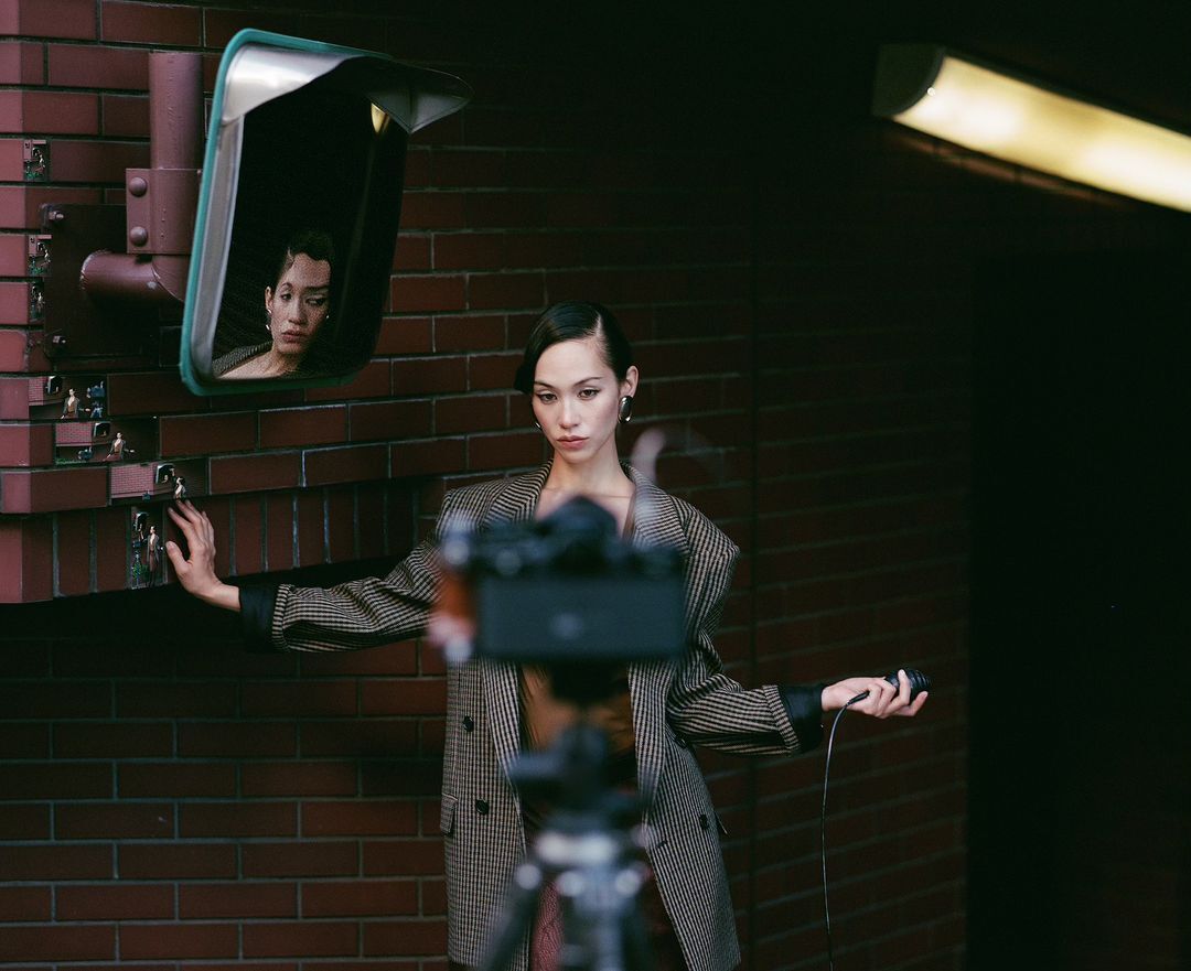 Young woman in a blazer posing near a brick wall with a mirror and lights, as a digital camera records the scene in soft, muted colors.