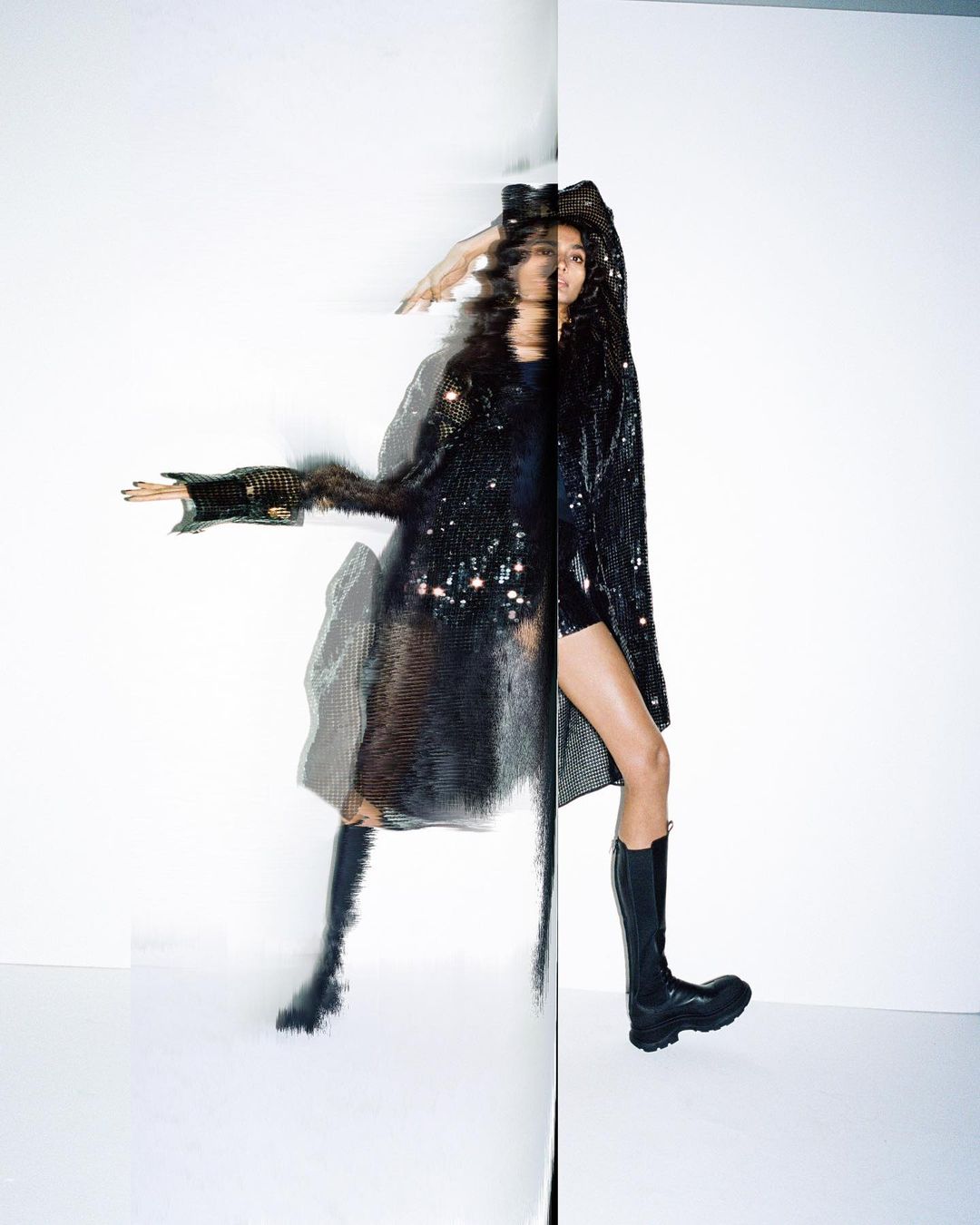 A slender woman in a sequined coat and black boots poses against a split white background, creating a mirrored effect.