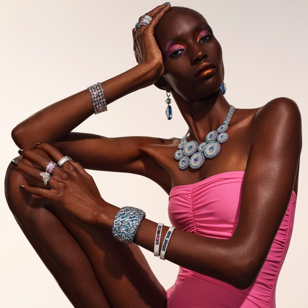 Dark-skinned woman poses with jewelry on white background, wearing a pink strapless top, bracelets, earrings, rings, and a necklace.