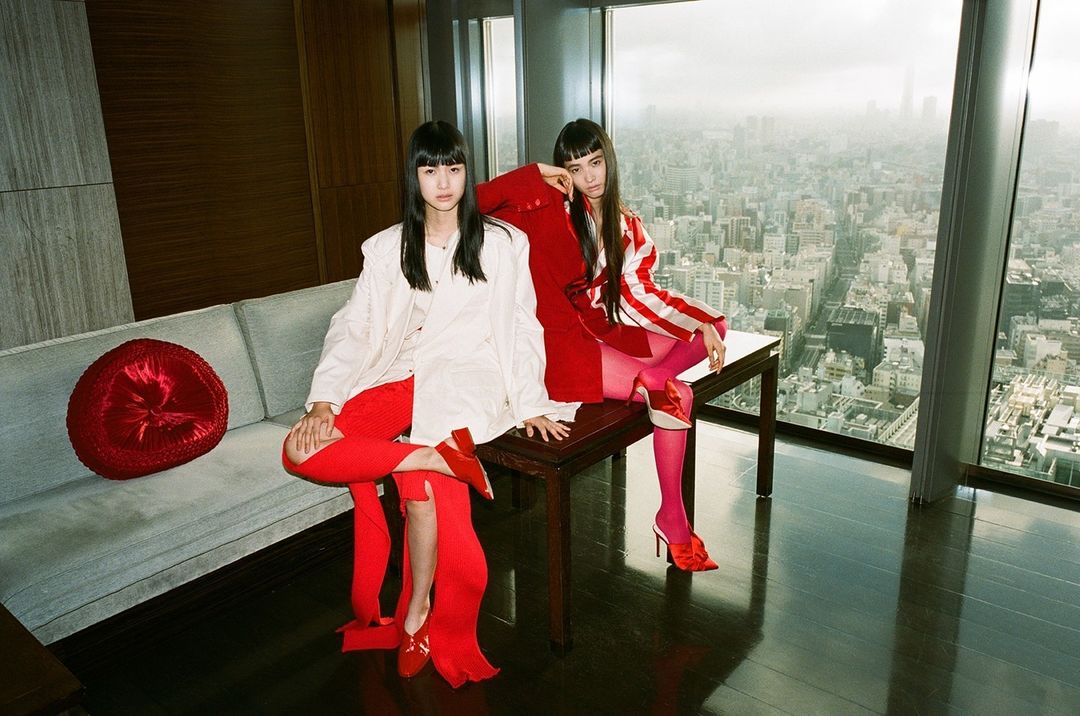 Two women in fashionable red and white outfits pose indoors against a cityscape backdrop. One sits on a grey sofa; the other, on a dark table.