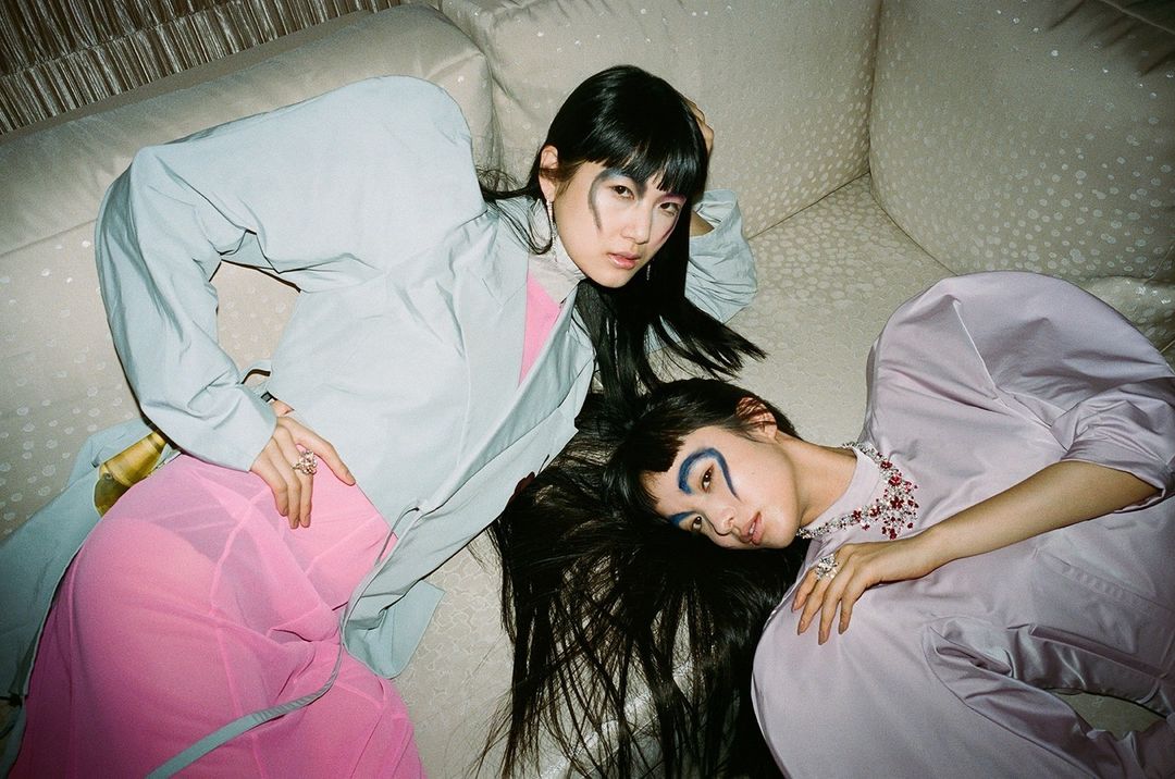 Two Asian women lie on a light patterned couch, wearing pastel jackets, with artistic makeup around their eyes, looking at the camera.