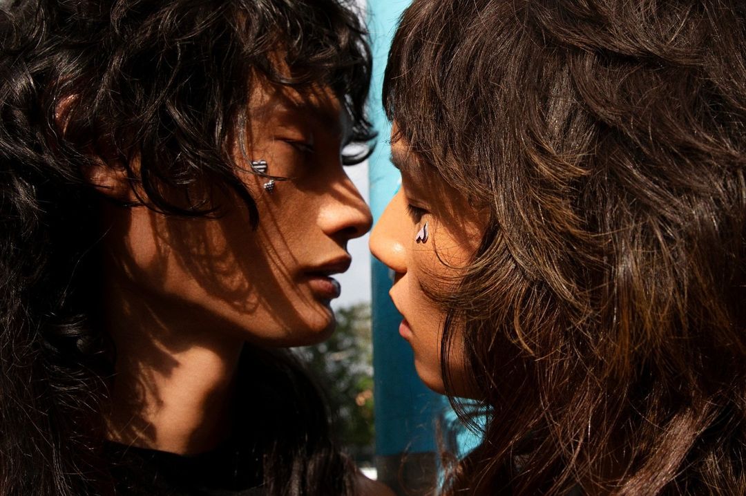 Close-up of two adult women facing each other with fair skin, wavy brown hair, and minimalist silver piercings against a blue background.