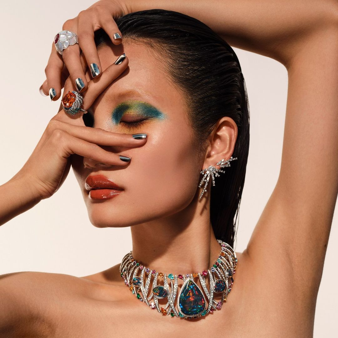 Close-up of a young Asian woman wearing eye-catching makeup and jewelry, including a necklace, earrings, and rings, on a white background.