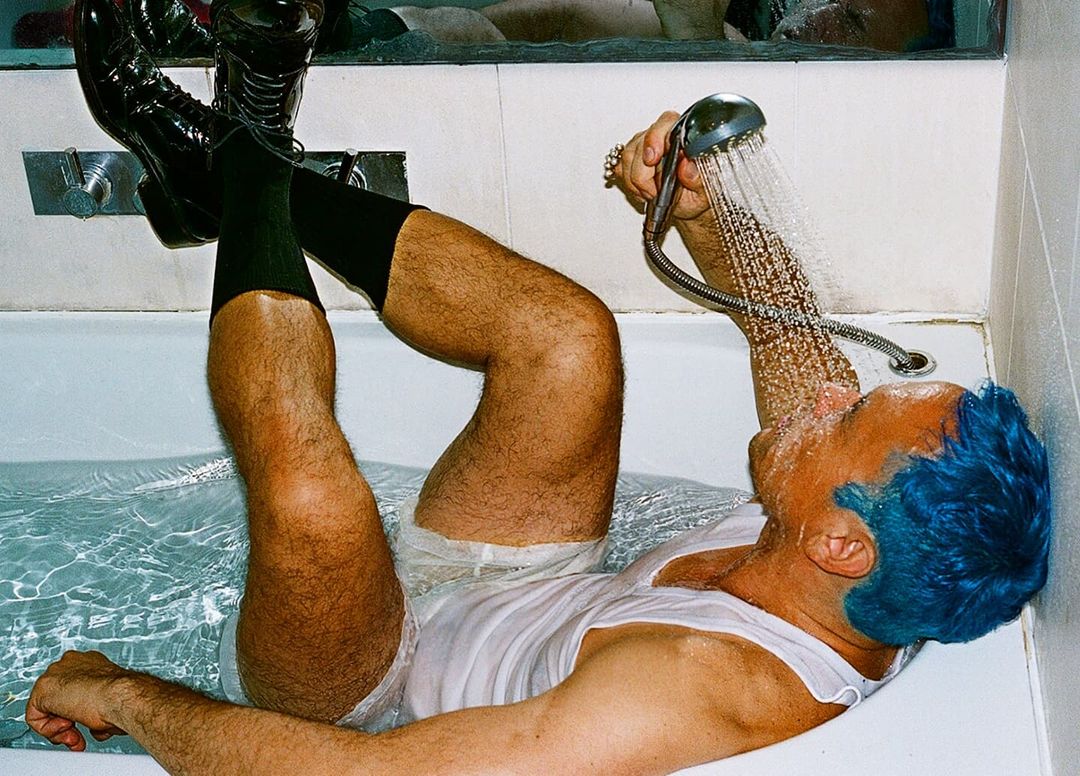 Adult male with blue hair lying in bathtub, legs raised, wearing boots and socks, showering his face with handheld shower head.