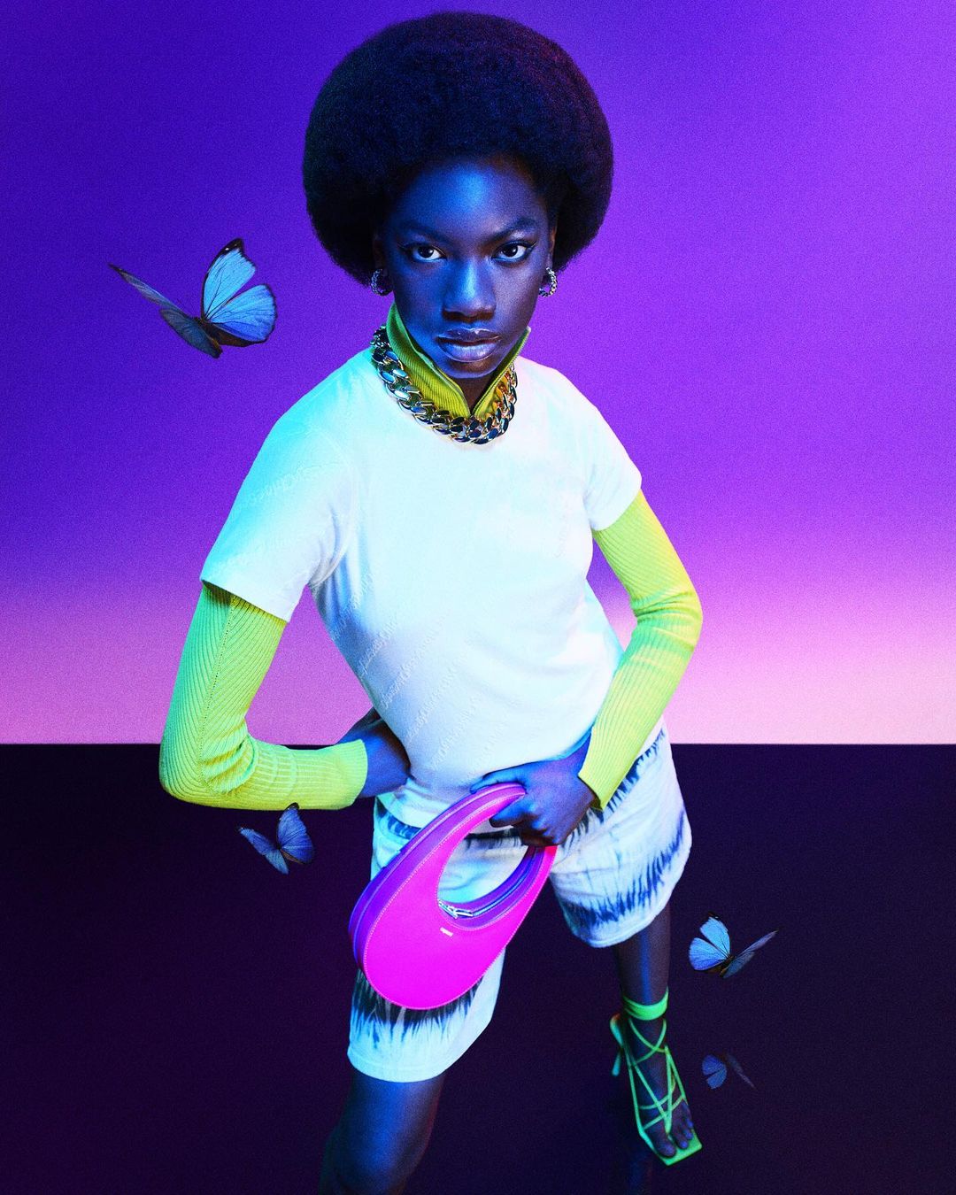 A dark-skinned woman with an afro, wearing a white t-shirt, tie-dye shorts, and neon green accessories, poses with a pink bag on a purple background.