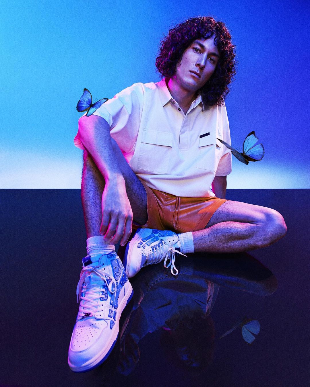 A young adult male with curly hair sits with legs crossed and is surrounded by blue butterflies, wearing a polo shirt, shorts, and sneakers.
