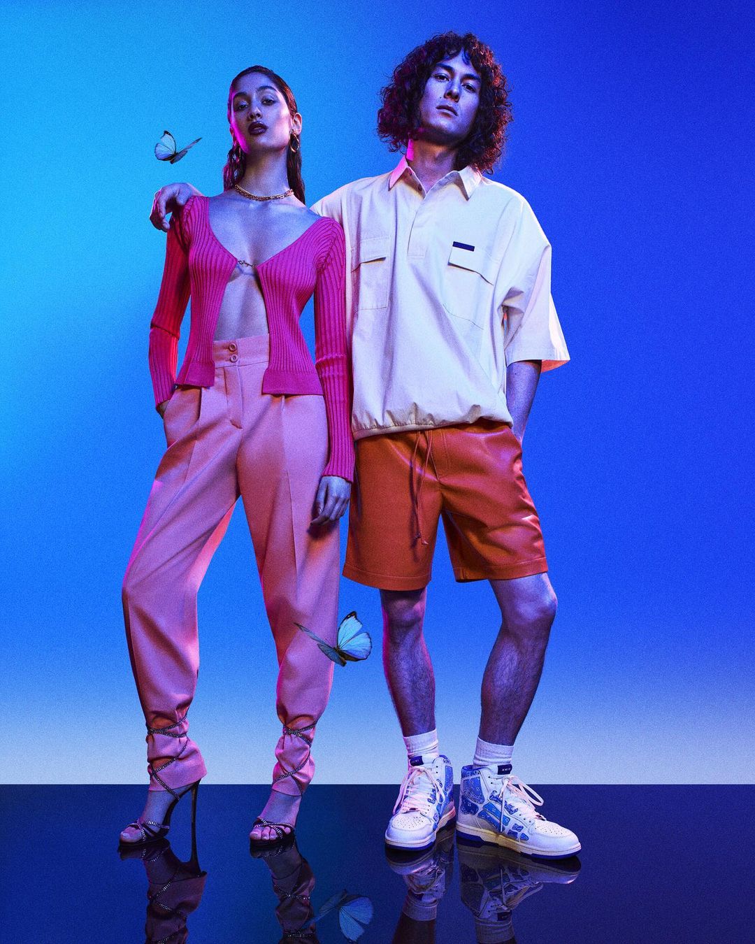 Two fashion models pose against a blue backdrop, wearing color-coordinated pink and orange outfits with sneakers. Butterflies added in post.