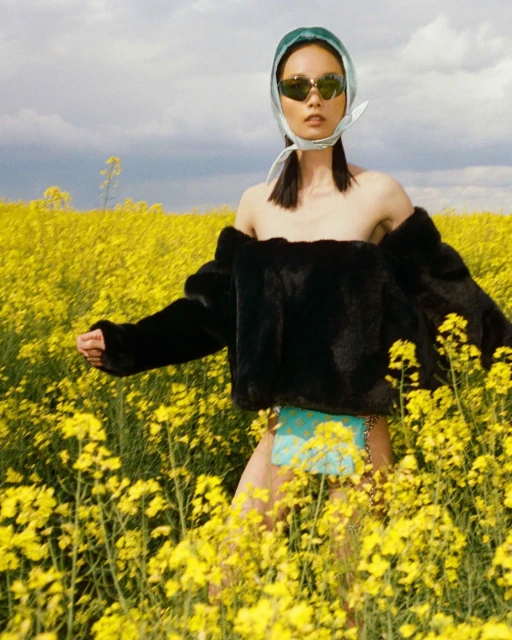 Asian woman in a field of yellow flowers wears a black fur jacket, sunglasses, and a headscarf in a fashion-style photograph.