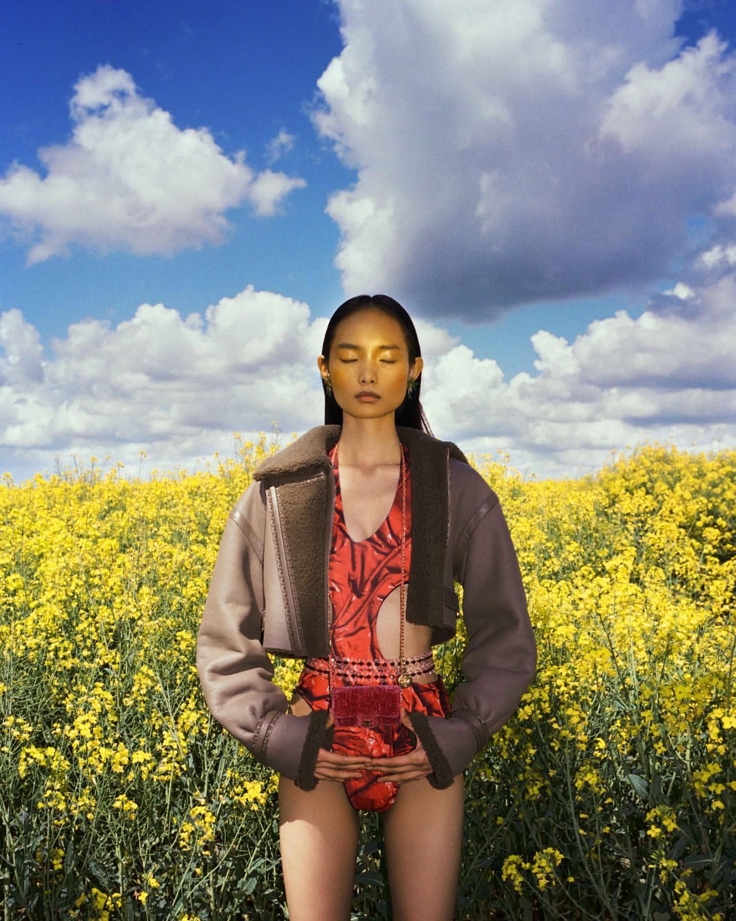 Woman in a red swimsuit and beige jacket stands in a yellow flower field under a blue sky with clouds in a fashion style portrait.