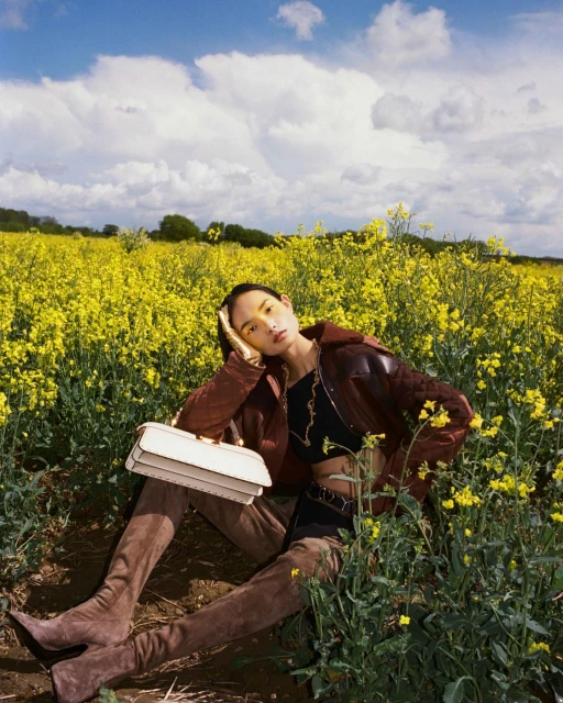 Woman in brown jacket and boots sits in field of yellow flowers with handbag, under blue sky with clouds. Fashion, outdoors.