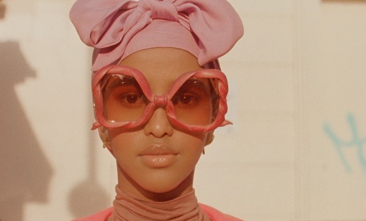 Close-up shot of a woman with a pink headwrap and oversized sunglasses against a textured wall with pale colors and some graffiti.