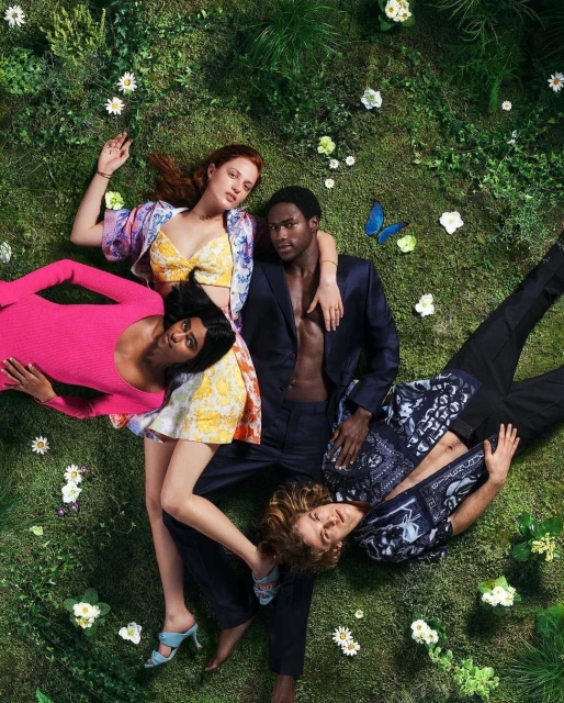 Four diverse adults lie in a field of grass and daisies, two women in dresses, one man in a suit, and one man in a patterned shirt.