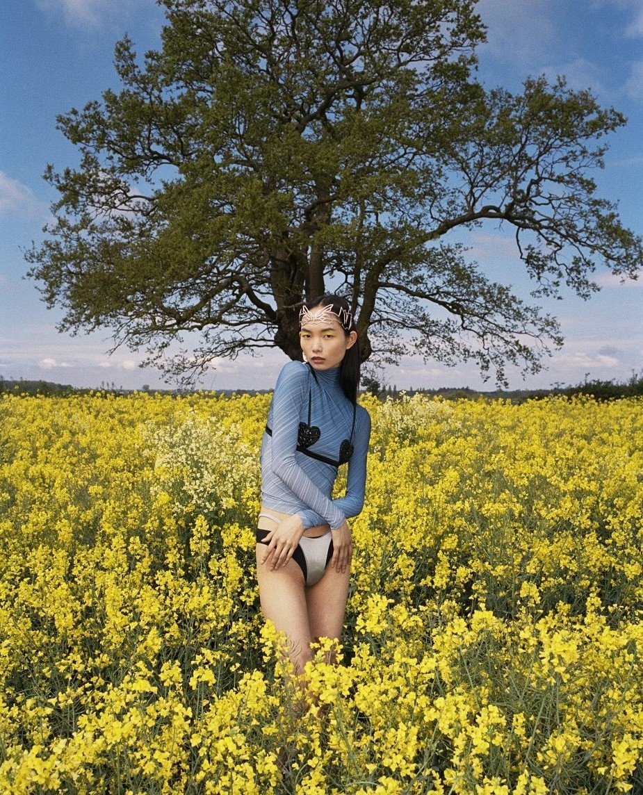 A woman stands in a field of yellow flowers under a blue sky, wearing a blue top, white bottoms, and a jeweled headpiece for a fashion shot.