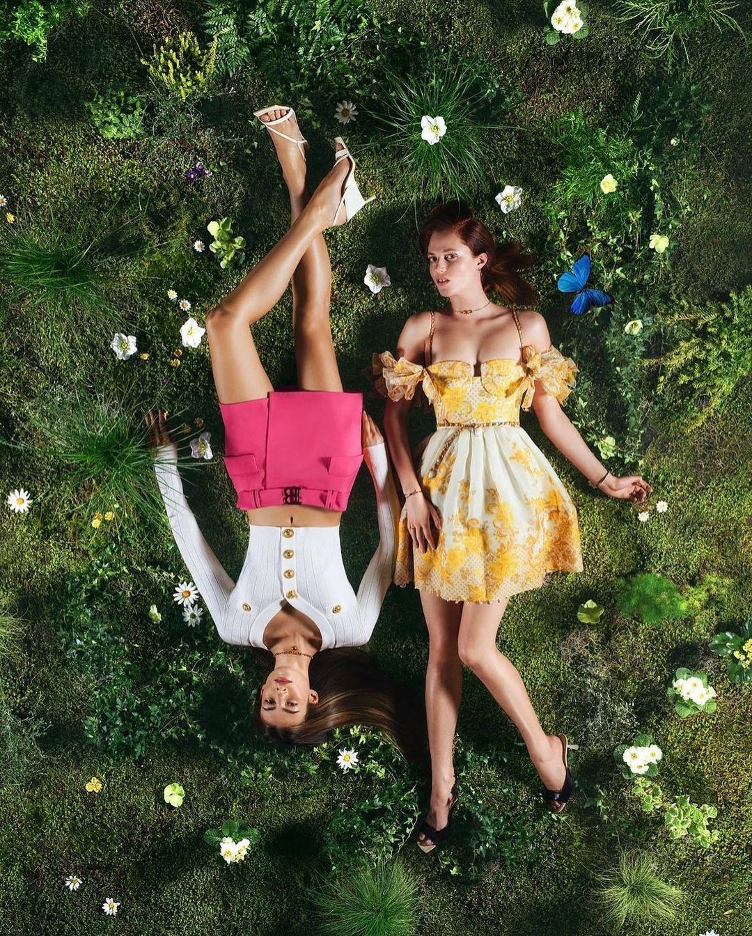 Two models lying on a lush green grassy field dotted with small white flowers and a blue butterfly.