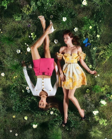 Two models lying on a lush green grassy field dotted with small white flowers and a blue butterfly.