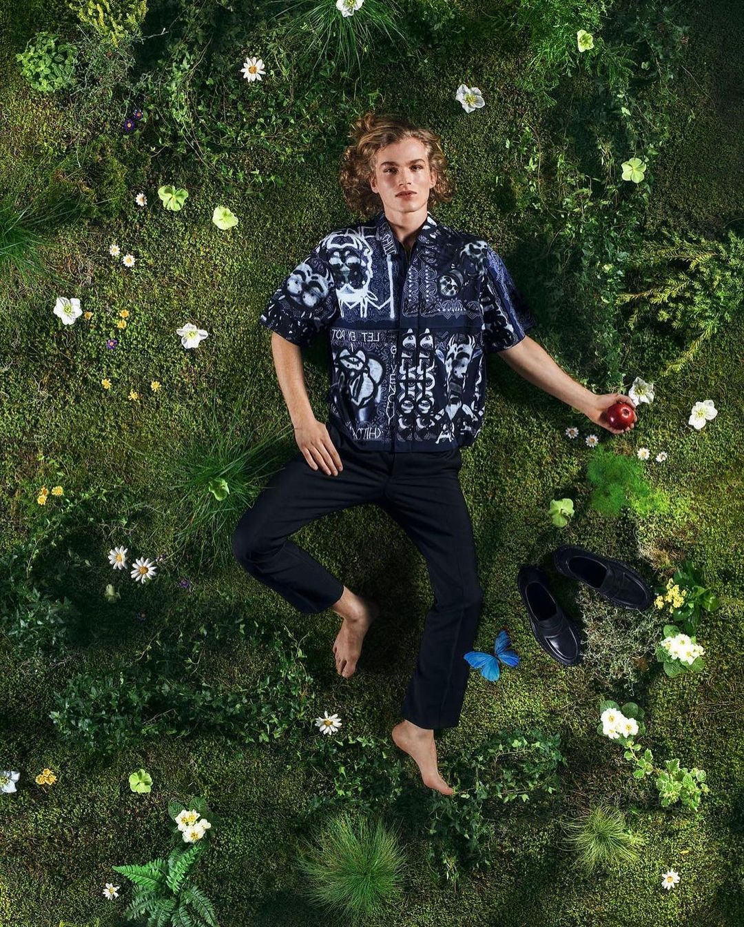 An aerial shot of a young man lying on a mossy surface with flowers, wearing a printed shirt and dark pants, holding an apple.