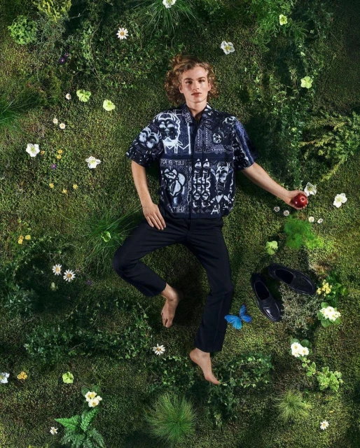 An aerial shot of a young man lying on a mossy surface with flowers, wearing a printed shirt and dark pants, holding an apple.