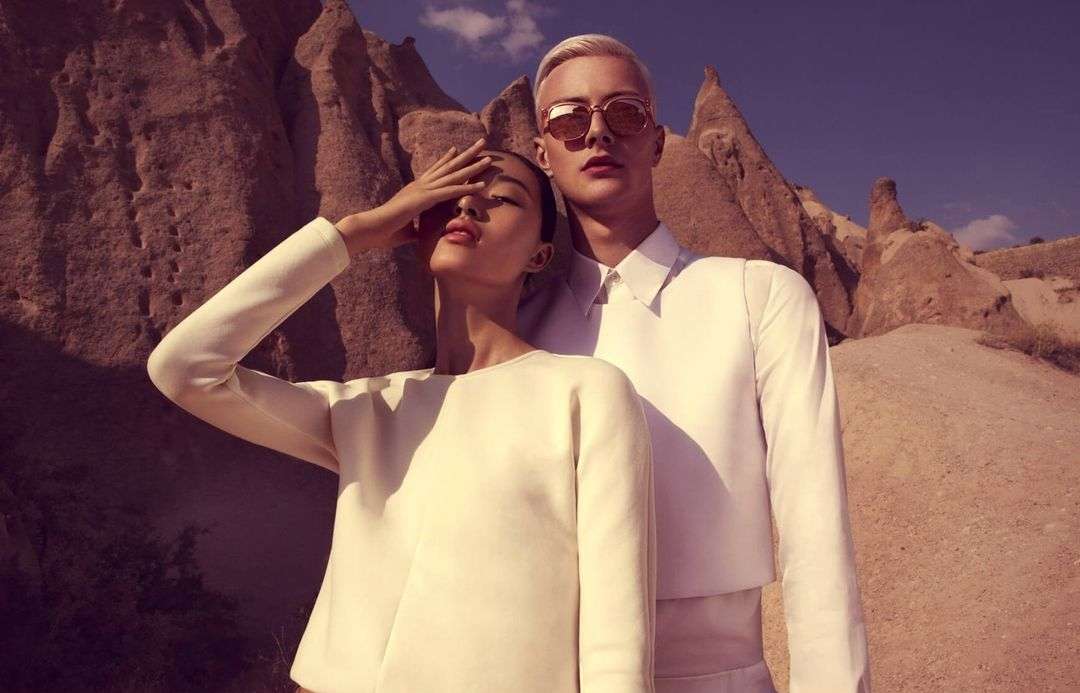 Fashion shot featuring a stylish couple in matching white outfits posing outdoors against a dramatic rock formation on a sunny day.
