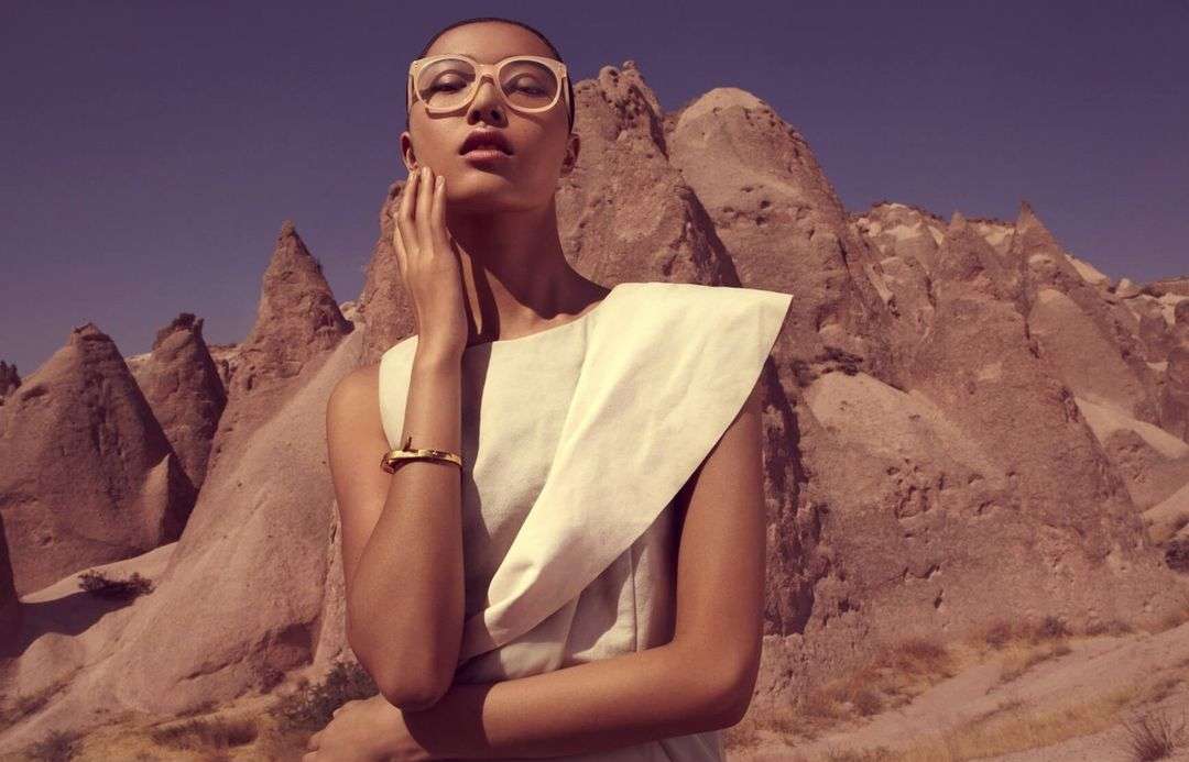 Outdoor fashion shot of a bald woman in a white dress and glasses posing against a backdrop of rock formations and a blue sky.