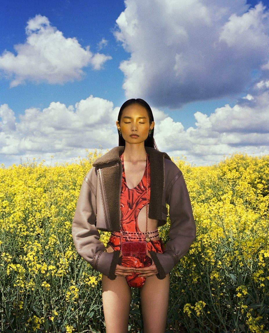 Woman in a patterned swimsuit and jacket stands in a field of yellow flowers under a blue, cloudy sky, eyes closed, holding a bag.